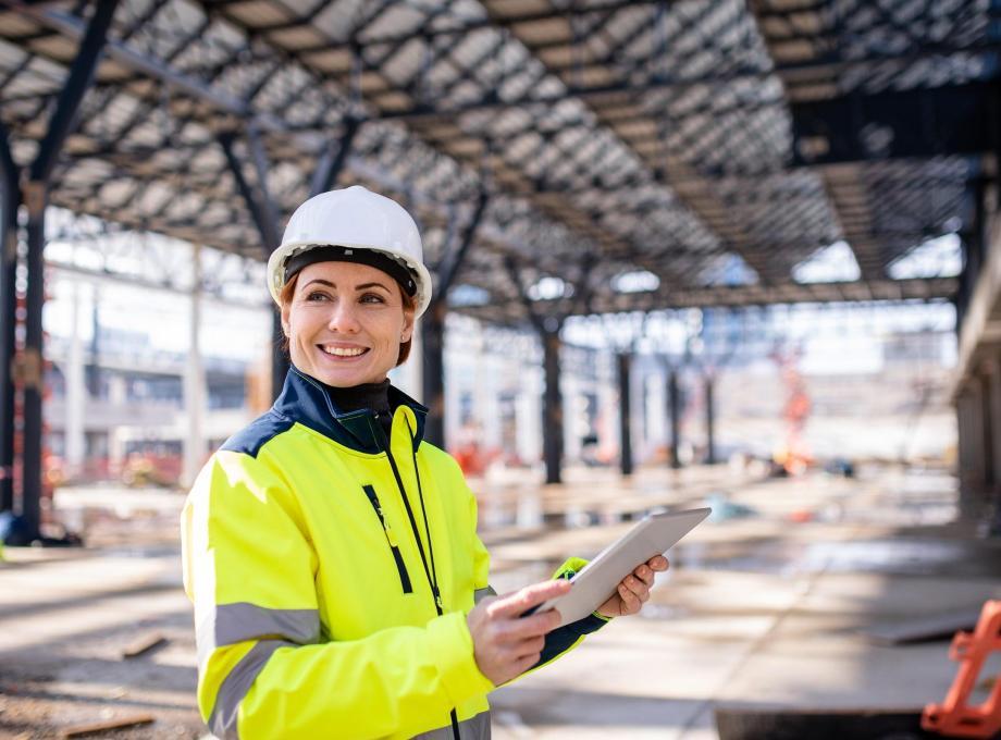 Femme souriante en gilet de sécurité et casque, tenant une tablette sur un chantier.