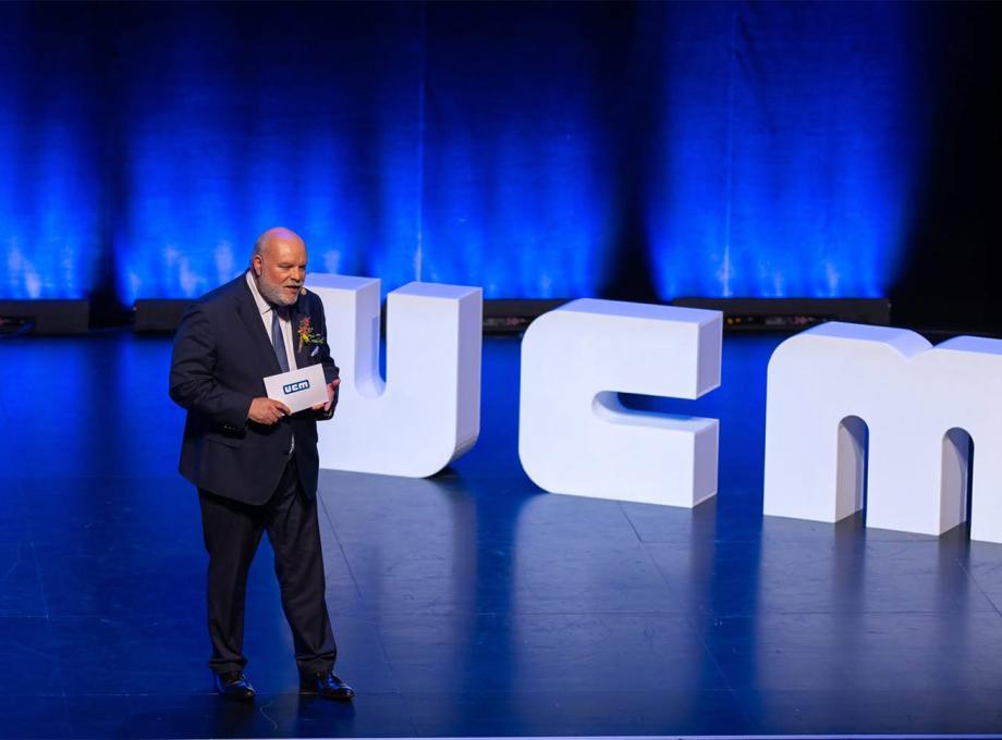 Un homme sur scène tenant un microphone, avec un fond lumineux bleu et le mot "UCM" en grandes lettres.